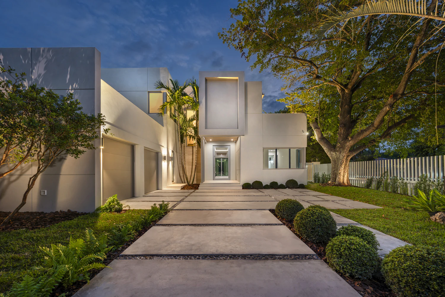 Modern home with a lit concrete path, clipped hedges and lawn at dusk