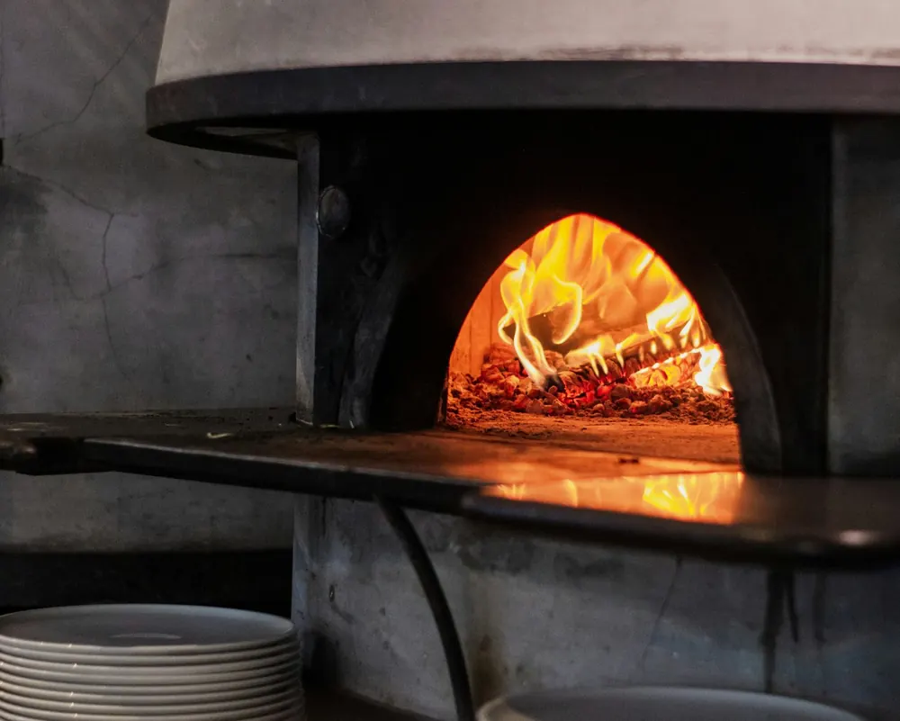 A pizza baking inside a stone woodfired oven with the fire glowing behind it