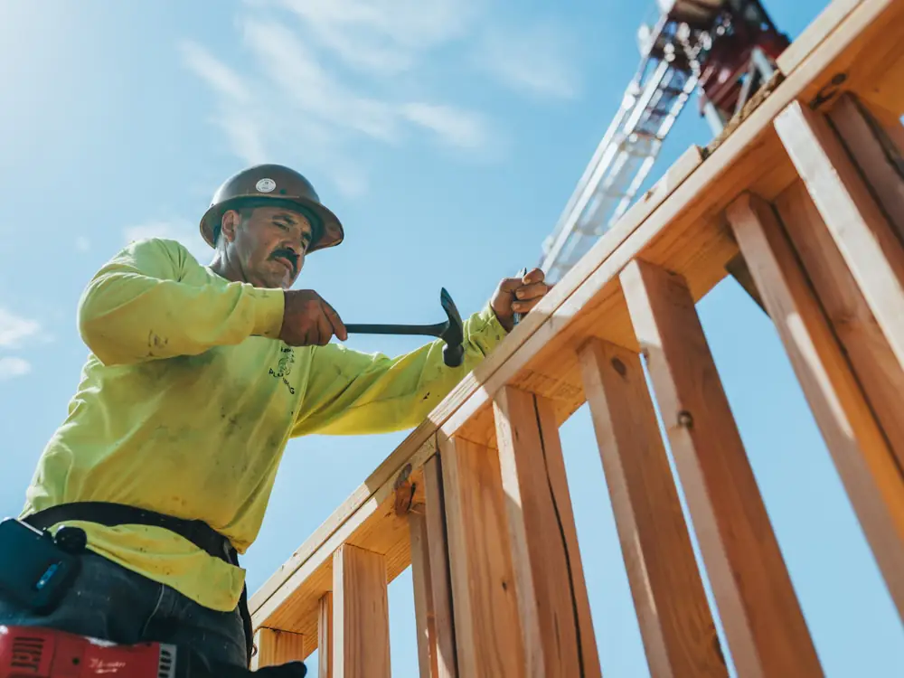 Carpenter in a hard hat nailing off a timber wall frame against a blue sky