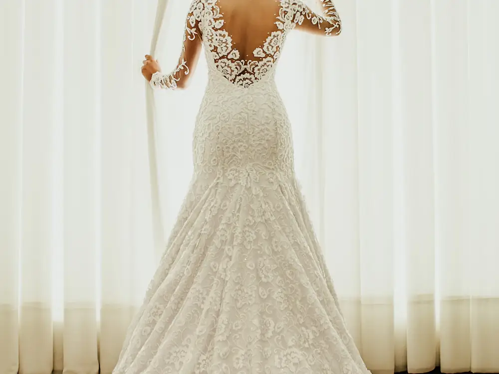 Back of a fitted lace gown with long lace sleeves and an illusion back, beside a sheer curtain