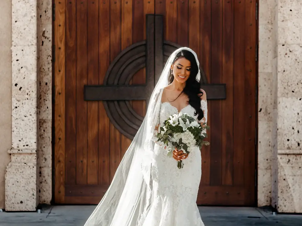 Bride in a lace A-line gown and cathedral veil, holding a white bouquet at a timber church door