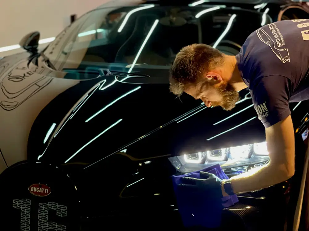 Detailer bent over a black supercar, wiping near the headlight under studio light bars