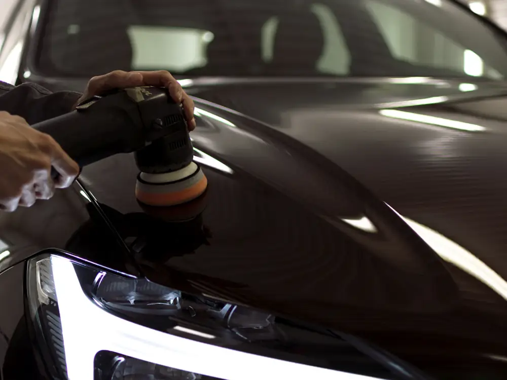 Machine polisher with an orange foam pad correcting black paint beside a headlight