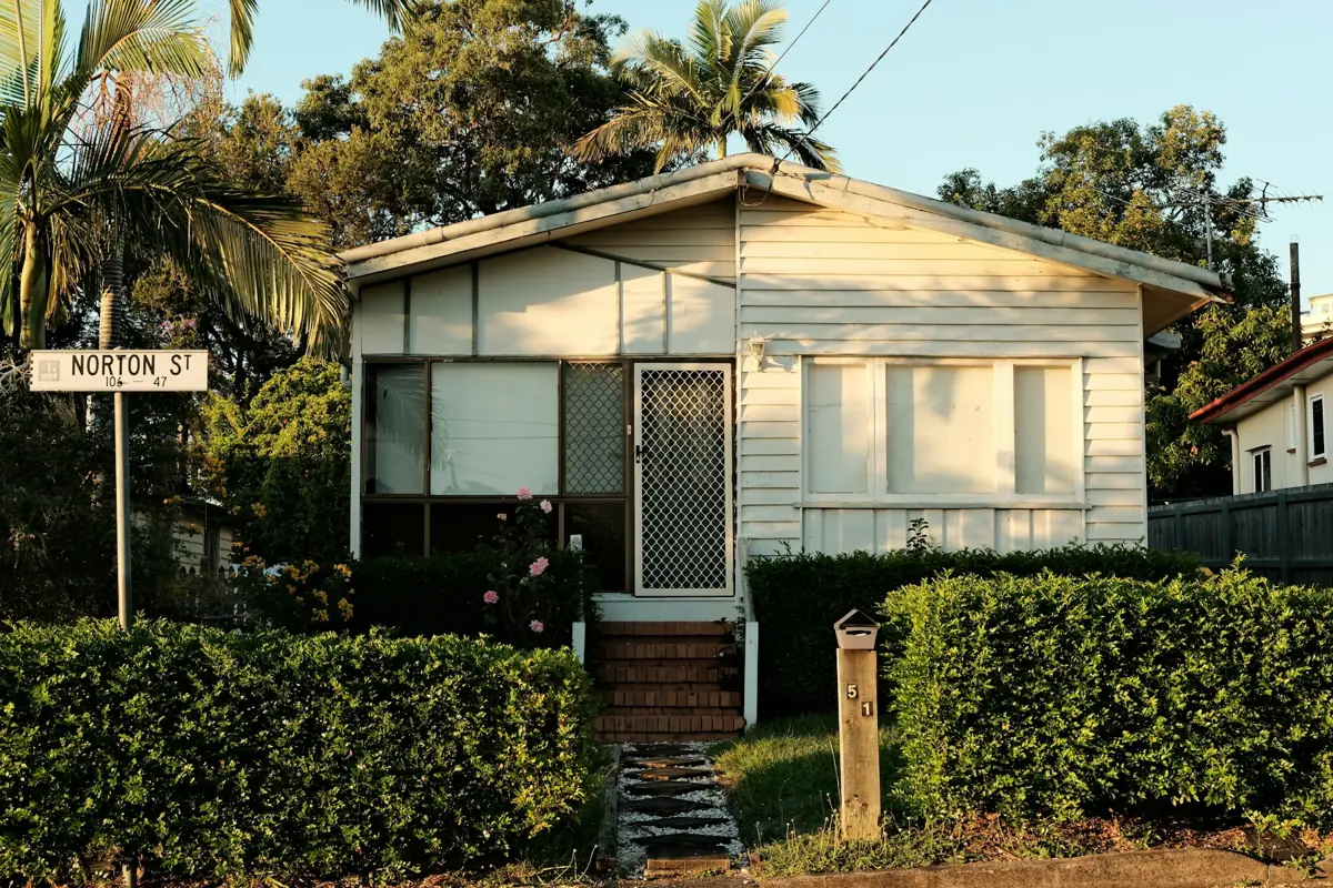 A low-set weatherboard house behind a hedge in a Brisbane street
