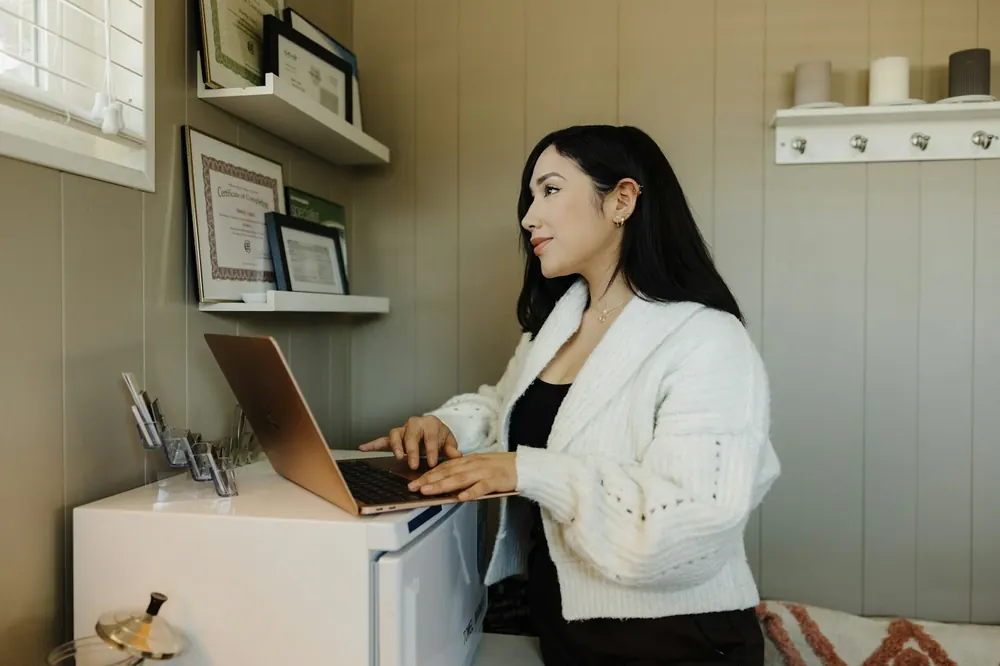 A bookkeeper working at a laptop in a small office with framed certificates on the shelf