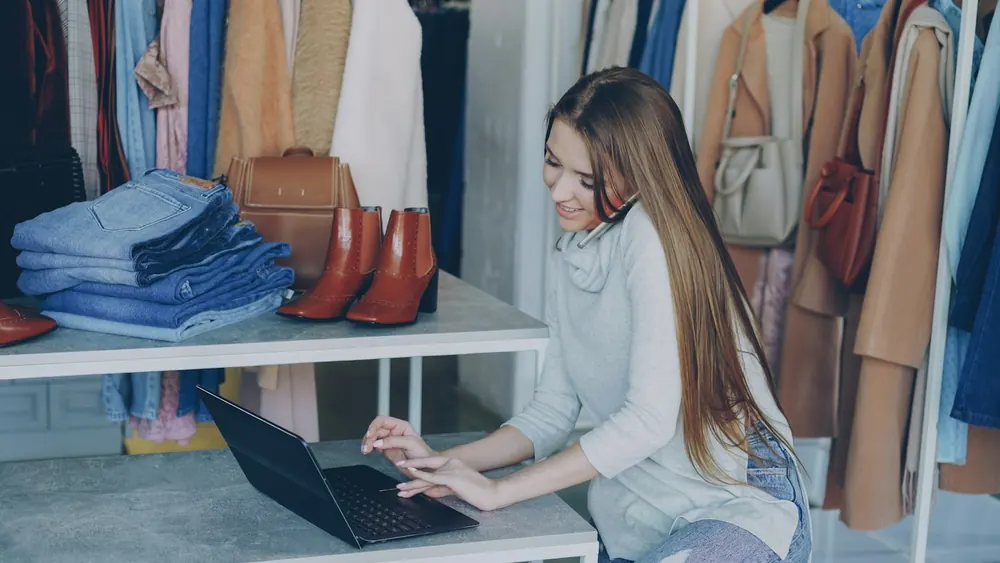 A boutique owner working on a laptop beside racks of clothes and a shelf of denim