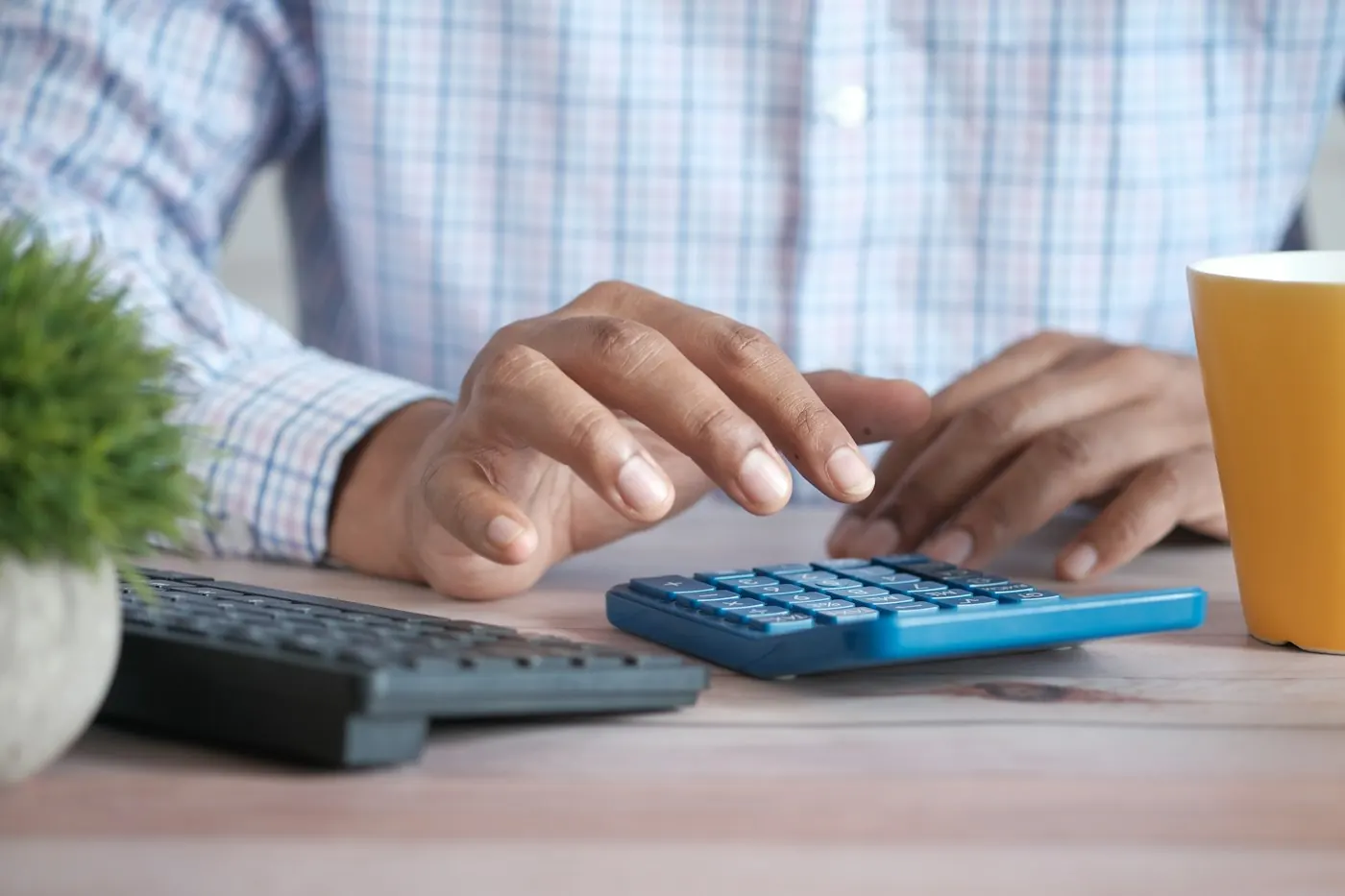 A bookkeeper's hands tapping a blue calculator beside a keyboard and a coffee mug