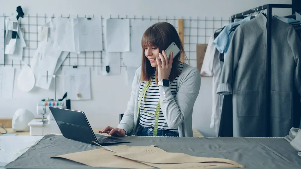 A designer smiling on the phone at her laptop in a bright workroom