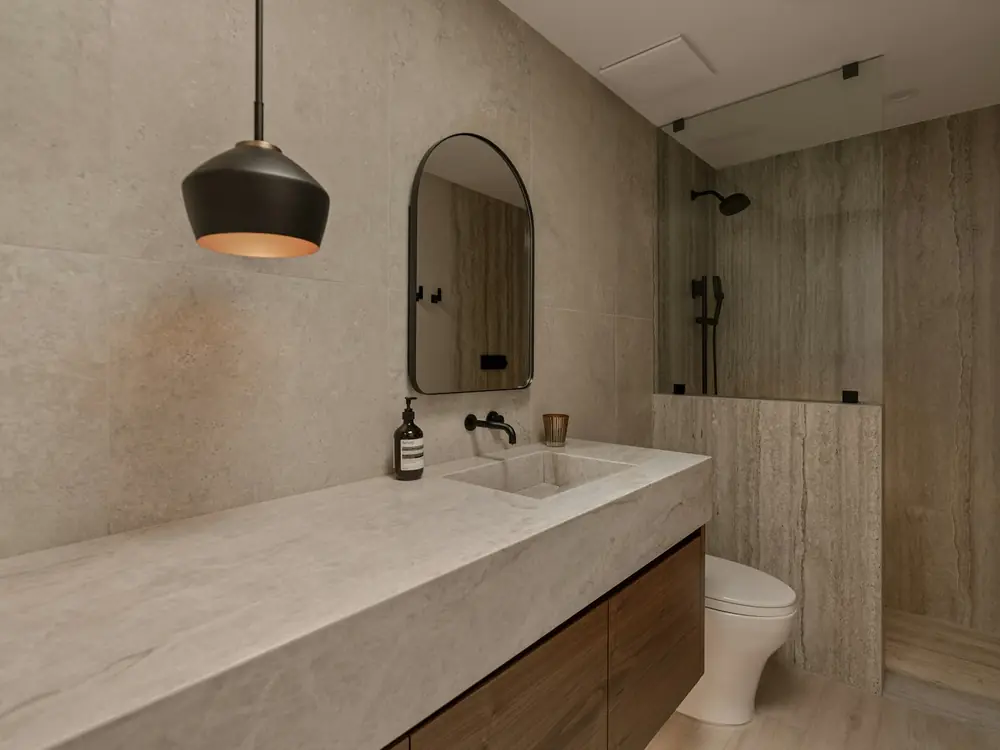 Bathroom in warm travertine-look stone with a long stone vanity and arched mirror