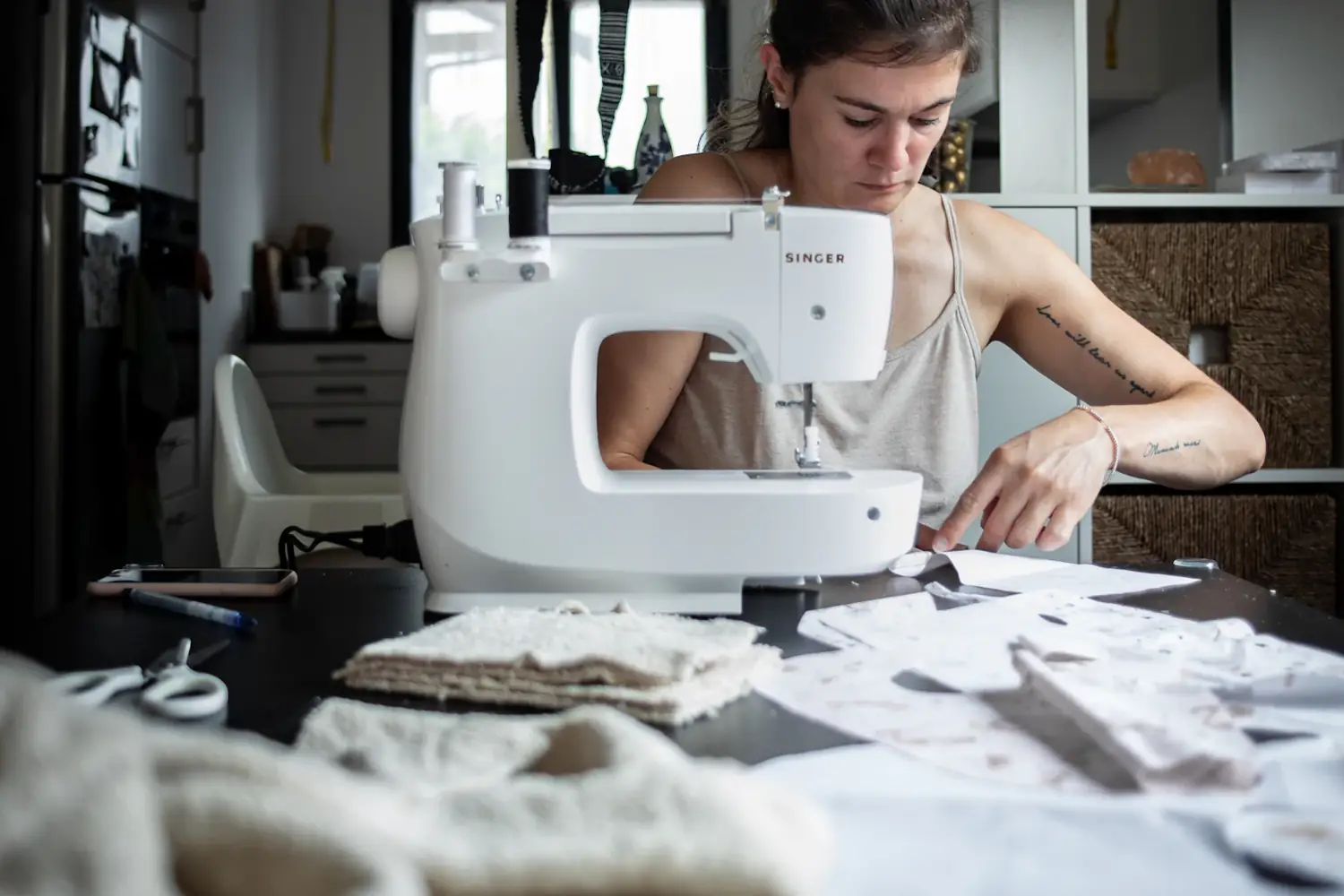 A dressmaker working at a white sewing machine in a bright studio, with fabric and patterns on the table