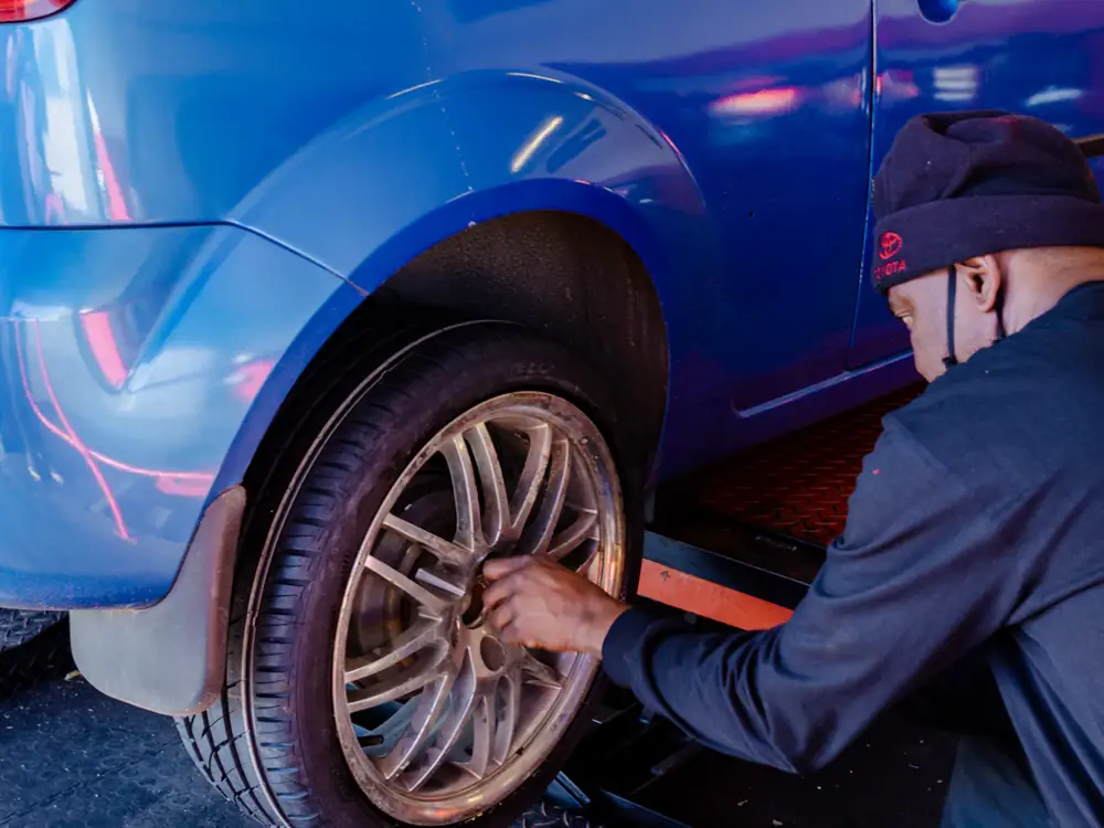 A technician kneeling beside a blue hatchback, working on the rear wheel